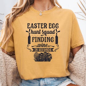 May include: Mustard-colored t-shirt with the text "EASTER EGG hunt squad FINDING wine IN SECONDS." The graphic includes wine bottles and Easter eggs. The shirt is a casual style.
