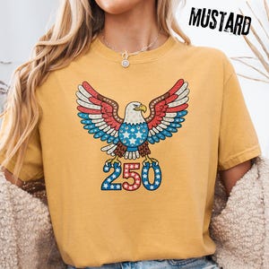 May include: Mustard-colored t-shirt featuring a patriotic eagle design with red, white, and blue accents. The eagle has outstretched wings and is above the number "250". The word "MUSTARD" is printed in black at the top right.