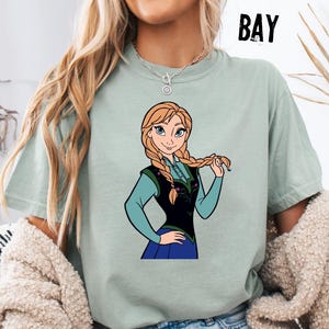 May include: A pale green t-shirt with a cartoon image of a character with blonde braids, wearing a black vest and blue dress. The word "BAY" is printed in black on the upper right side of the shirt.