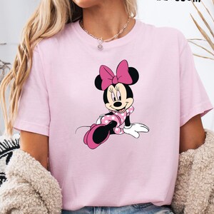 May include: A light pink t-shirt featuring a Minnie Mouse graphic. The cartoon character is depicted in a pink polka-dotted dress and bow. The word "BLOSSOM" is printed in black at the top right.