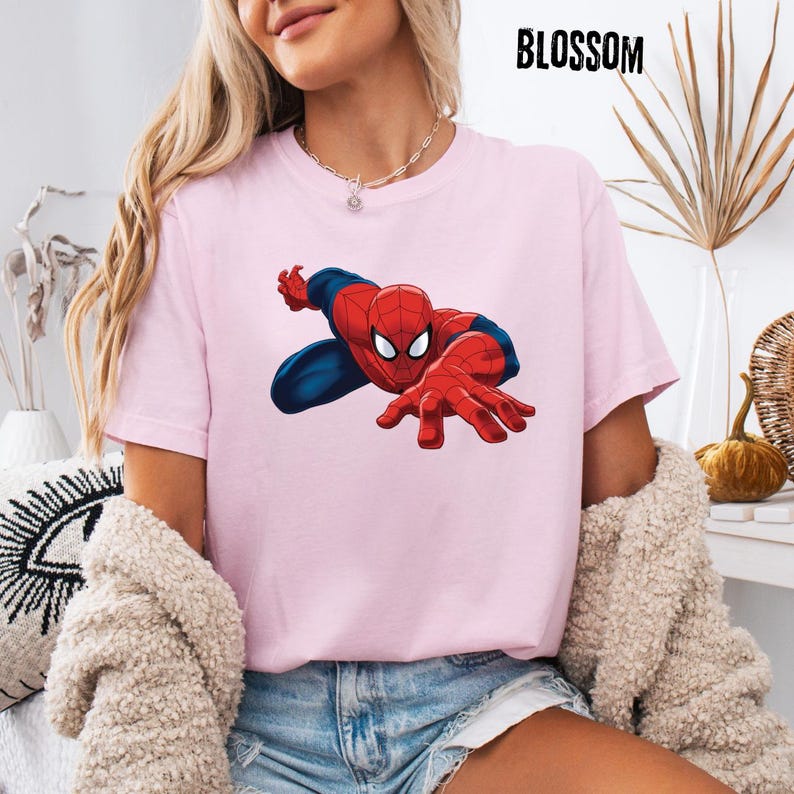 May include: A light pink short-sleeved t-shirt featuring a graphic of the character Spider-Man in a crouching pose with red and blue colors. The word BLOSSOM is printed in black capital letters in the upper right corner of the image.