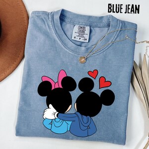 May include: Blue jean colored t-shirt featuring a graphic of Mickey and Minnie Mouse embracing, with red hearts above them. The shirt has a crew neck and short sleeves. The text "BLUE JEAN" is at the top of the image.