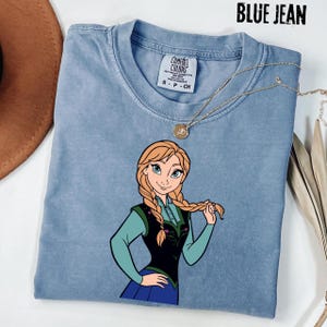 May include: A blue jean Comfort Colors t-shirt with a cartoon illustration of a character with blonde braids, wearing a green and black outfit. A gold necklace with a pendant is draped on the shirt. The text "BLUE JEAN" is at the top.
