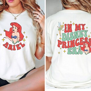 May include: A cream-colored t-shirt featuring a graphic of Ariel on the front with the text ARIEL below. The back of the shirt displays a graphic of Tinker Bell and Ariel with the text IN MY DISNEY PRINCESS ERA surrounded by yellow stars.