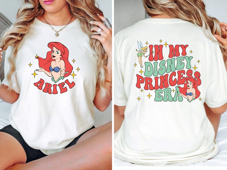 May include: A cream-colored t-shirt featuring a graphic of Ariel on the front with the text ARIEL below. The back of the shirt displays a graphic of Tinker Bell and Ariel with the text IN MY DISNEY PRINCESS ERA surrounded by yellow stars.