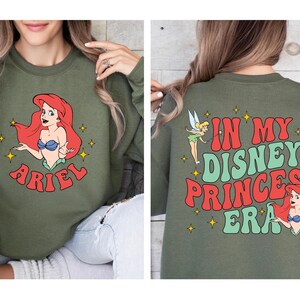 May include: An olive green crewneck sweatshirt featuring a graphic of Ariel on the front with the name Ariel below. The back displays Tinker Bell and Ariel with the text In My Disney Princess Era in red and green, accented by yellow stars.