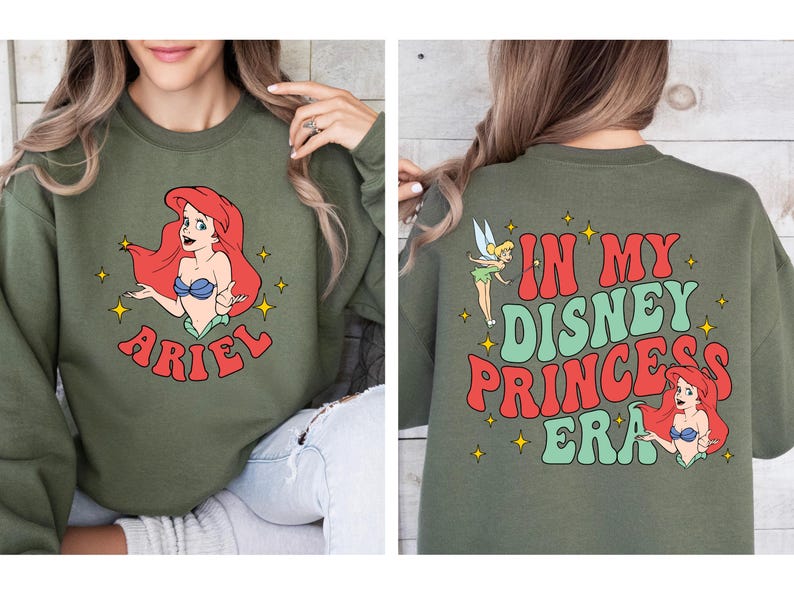 May include: An olive green crewneck sweatshirt featuring a graphic of Ariel on the front with the name Ariel below. The back displays Tinker Bell and Ariel with the text In My Disney Princess Era in red and green, accented by yellow stars.