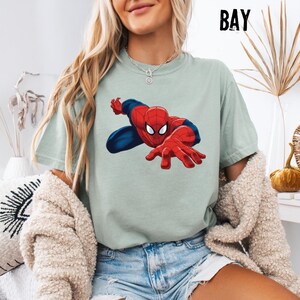 May include: A sage green short-sleeve t-shirt featuring a graphic print of the superhero Spider-Man in a dynamic crouching pose with red and blue colors. The shirt has a crew neckline and is worn with blue denim shorts and a textured beige cardigan.