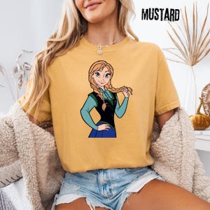May include: Mustard-coloured t-shirt with a cartoon illustration of a character with blonde braids, wearing a blue and black outfit. The word "MUSTARD" is printed in black, upper-case letters in the top right corner. The shirt is worn by a person.