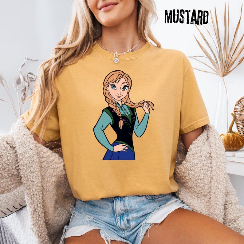 May include: Mustard-coloured t-shirt with a cartoon illustration of a character with blonde braids, wearing a blue and black outfit. The word "MUSTARD" is printed in black, upper-case letters in the top right corner. The shirt is worn by a person.
