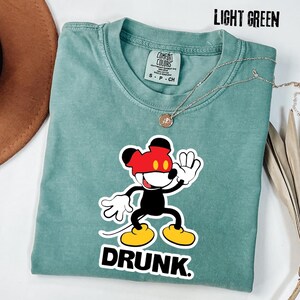 May include: Light green t-shirt featuring a cartoon mouse character with a red mask and yellow shoes. The word "DRUNK" is printed in bold white letters below the character. A silver necklace with a pendant is also visible.