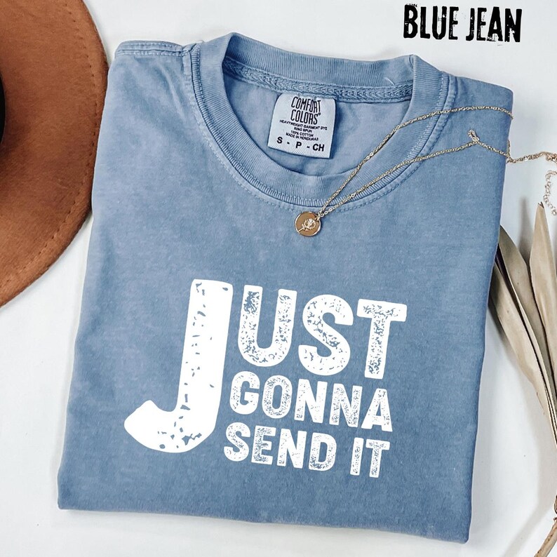 May include: Blue jean colored t-shirt with the text "JUST GONNA SEND IT" in white block letters. The shirt has a crew neck and short sleeves. A gold necklace with a pendant is draped across the shirt.