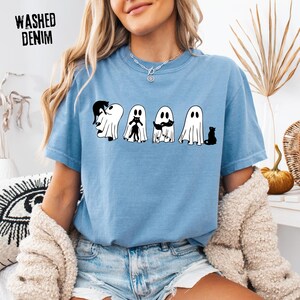 Pu&ograve; includere: Una t-shirt azzurra con una grafica di quattro fantasmi bianchi che interagiscono con gatti neri. Il testo WASHED DENIM &egrave; stampato nell'angolo in alto a sinistra. I fantasmi hanno semplici occhi e bocche ovali e gatti neri vicino a loro.