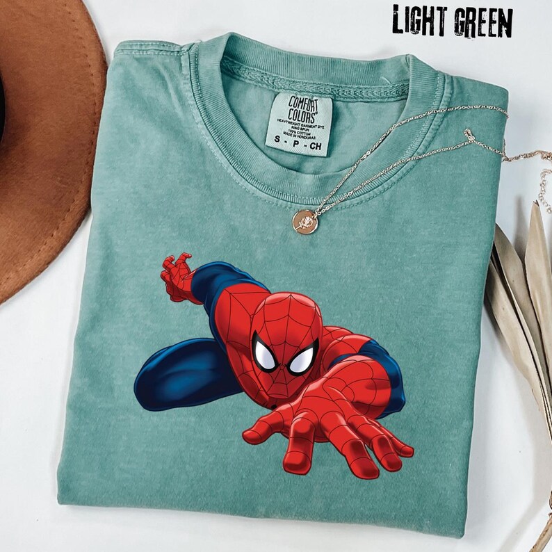 May include: A light green short-sleeve t-shirt featuring a graphic of the character Spider-Man in a crouching pose. The shirt has a crew neckline and a Comfort Colors brand label at the inner collar. The text LIGHT GREEN is visible at the top.