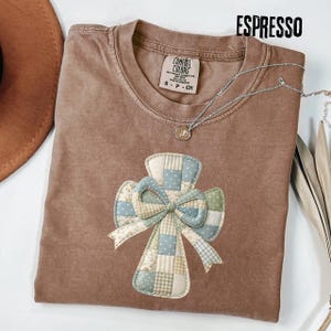 May include: Espresso-coloured t-shirt featuring a patchwork cross design in blue, green, and cream. The shirt has a crew neck and a small tag near the collar. A silver necklace with a pendant is draped across the shirt.
