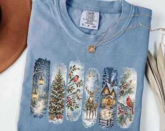 Può includere: Una t-shirt color blu jeans con etichetta Comfort Colors sul collo. Il davanti presenta sei pannelli rettangolari verticali che raffigurano scene invernali, tra cui un albero di Natale illuminato, un cervo, uccelli e una baita, con una collana dorata.