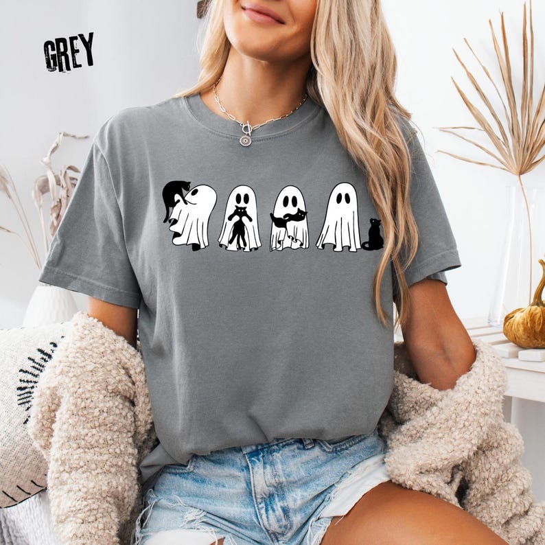 Pu&ograve; includere: Una t-shirt grigia a maniche corte con una stampa grafica di quattro fantasmi bianchi che interagiscono con gatti neri. Il testo GREY &egrave; stampato nell'angolo in alto a sinistra. Il design mostra fantasmi che tengono, abbracciano e siedono accanto a gatti.