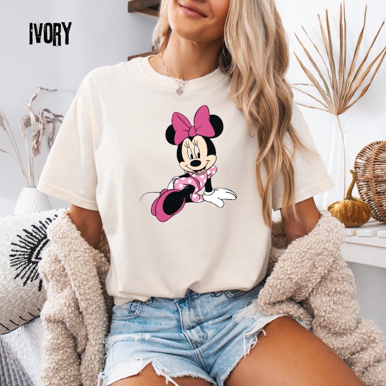 May include: Ivory-colored t-shirt featuring a graphic of Minnie Mouse in a pink polka-dot dress and bow. The word "IVORY" is printed in black at the top left. The shirt is a casual, short-sleeved style.