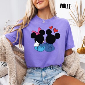 May include: A violet t-shirt featuring a graphic of two cartoon mice embracing, with red heart accents. The text "VIOLET" is printed in black at the top right. The shirt is a comfortable, casual style.