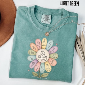 May include: Light green t-shirt with a floral design. The flower design has colorful petals with positive affirmations and the text "YOU CAN BLOOM every day". A silver necklace with a pendant is on the shirt.
