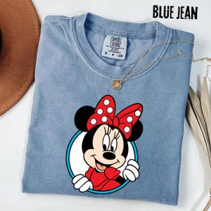 May include: Blue jean colored t-shirt featuring a Minnie Mouse graphic. The graphic shows Minnie Mouse with a red bow with white polka dots, a red dress, and white gloves. The text "BLUE JEAN" is at the top.