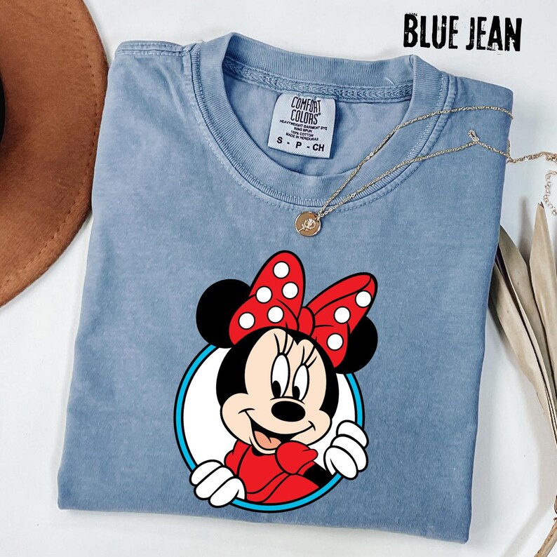 May include: Blue jean colored t-shirt featuring a Minnie Mouse graphic. The graphic shows Minnie Mouse with a red bow with white polka dots, a red dress, and white gloves. The text "BLUE JEAN" is at the top.