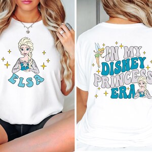 May include: A white t-shirt featuring a graphic of Elsa on the front with the name Elsa below. The back of the shirt displays Tinker Bell and Elsa with the text In My Disney Princess Era, surrounded by small yellow star icons throughout the design.