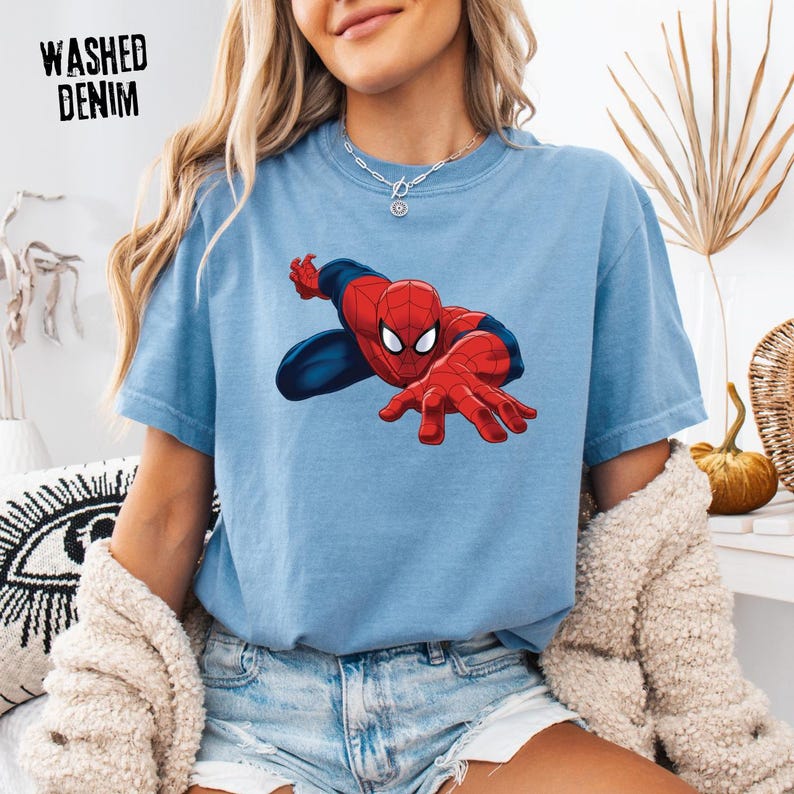 May include: A light blue washed denim t-shirt featuring a graphic of the superhero Spider-Man in a dynamic crouching pose with red and blue colors. The text WASHED DENIM is printed in black in the upper left corner of the shirt design.