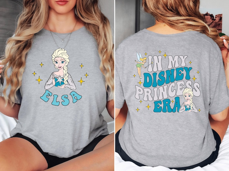 May include: A heather gray t-shirt featuring a graphic of Elsa on the front with the name Elsa below. The back of the shirt displays a graphic of Tinker Bell and Elsa with the text In My Disney Princess Era surrounded by yellow star icons.