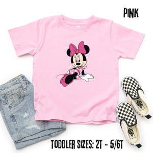 May include: Pink toddler t-shirt featuring a Minnie Mouse graphic. The shirt has a crew neck and short sleeves. The image also shows denim shorts and a pair of black and white checkered slip-on shoes. Text on the shirt reads "PINK" and "TODDLER SIZES: 2T - 5/6T".
