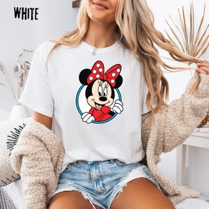May include: White t-shirt featuring a cartoon Minnie Mouse design. Minnie Mouse is wearing a red dress and a red bow with white polka dots. The design is framed by a blue circle. The word "WHITE" is printed in black at the top left.