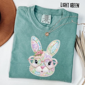 以下が含まれることがあります： ハート型のメガネをかけた、花のウサギのデザインのライトグリーンのTシャツ。ウサギはピンクと紫のアクセントの花柄です。シャツの上部には「LIGHT GREEN」の文字が入っています。