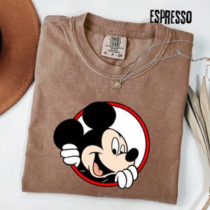 Pu&ograve; includere: T-shirt color espresso con grafica di Topolino che sbuca da un cerchio bordato di rosso. La maglietta ha girocollo e maniche corte. Una collana d'argento con un piccolo ciondolo &egrave; drappeggiata sulla maglietta.