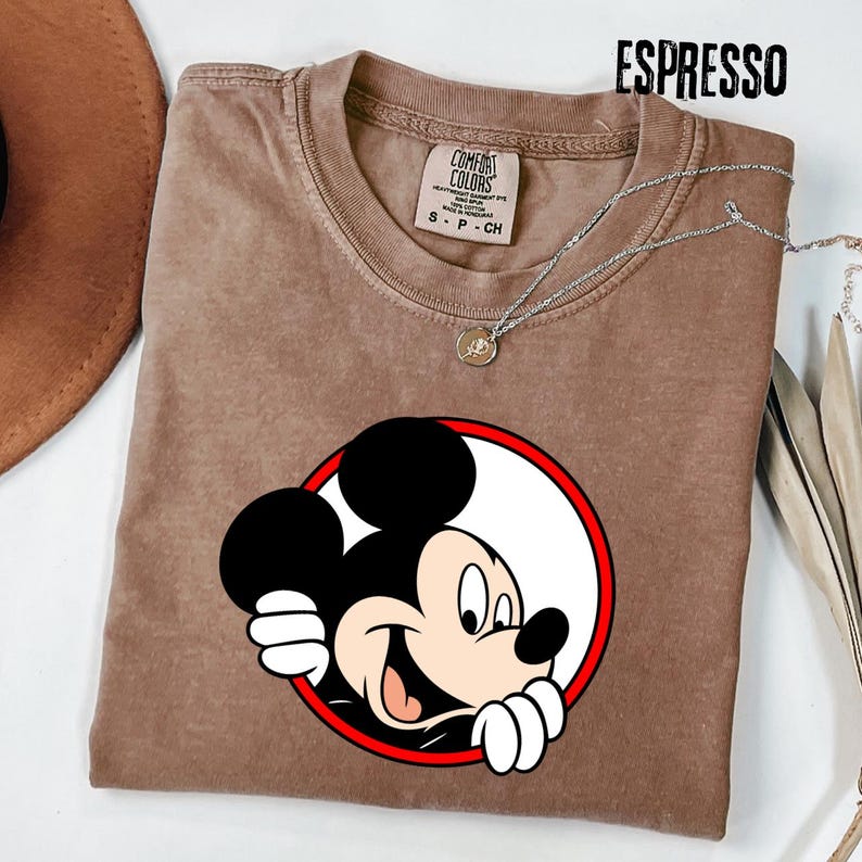 Pu&ograve; includere: T-shirt color espresso con grafica di Topolino che sbuca da un cerchio bordato di rosso. La maglietta ha girocollo e maniche corte. Una collana d'argento con un piccolo ciondolo &egrave; drappeggiata sulla maglietta.