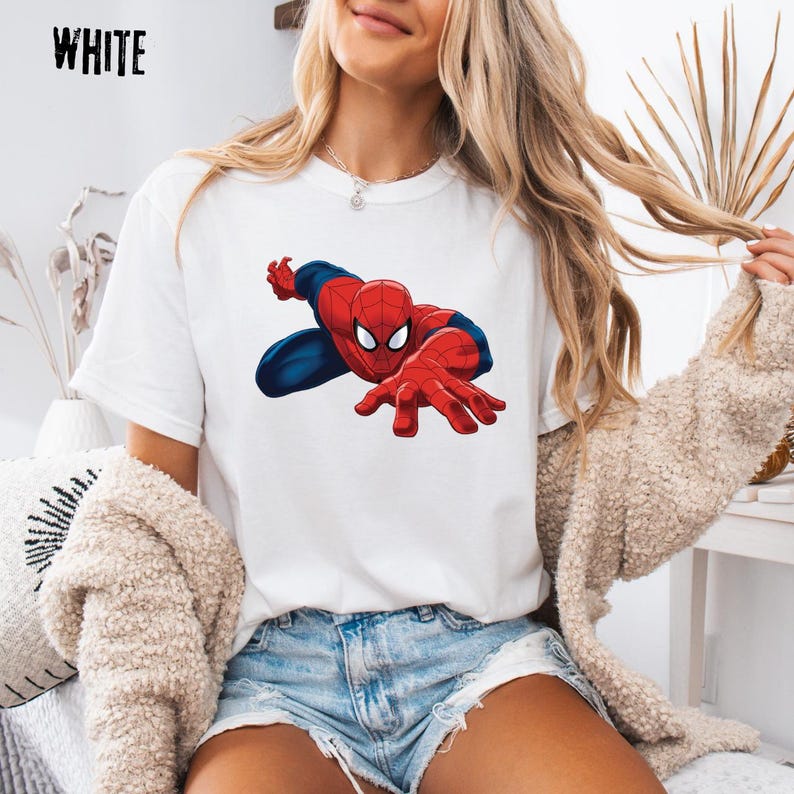 May include: A white short-sleeved t-shirt featuring a graphic print of the character Spider-Man in a dynamic crouching pose with red and blue colors. The word WHITE is printed in black text in the upper left corner of the image.
