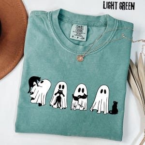 Pu&ograve; includere: Una t-shirt verde chiaro con una grafica raffigurante quattro fantasmi bianchi che interagiscono con gatti neri. La maglia presenta un girocollo e un'etichetta del marchio Comfort Colors all'interno. Il testo LIGHT GREEN &egrave; stampato nell'angolo in alto a destra.