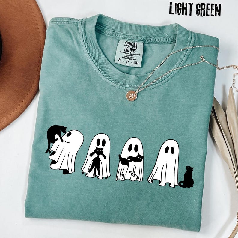 Pu&ograve; includere: Una t-shirt verde chiaro con una grafica raffigurante quattro fantasmi bianchi che interagiscono con gatti neri. La maglia presenta un girocollo e un'etichetta del marchio Comfort Colors all'interno. Il testo LIGHT GREEN &egrave; stampato nell'angolo in alto a destra.