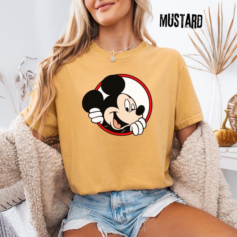 Pu&ograve; includere: T-shirt color senape con grafica di Topolino sorridente all'interno di un cerchio rosso e bianco. La parola "MUSTARD" &egrave; stampata in lettere nere in alto a destra. La maglietta &egrave; a maniche corte e girocollo.