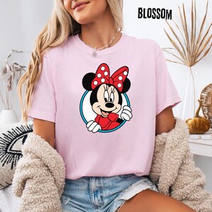 May include: A light pink t-shirt featuring a Minnie Mouse graphic. The cartoon character is framed in a blue and white circle, wearing a red bow with white polka dots. The word "BLOSSOM" is printed in black at the top.