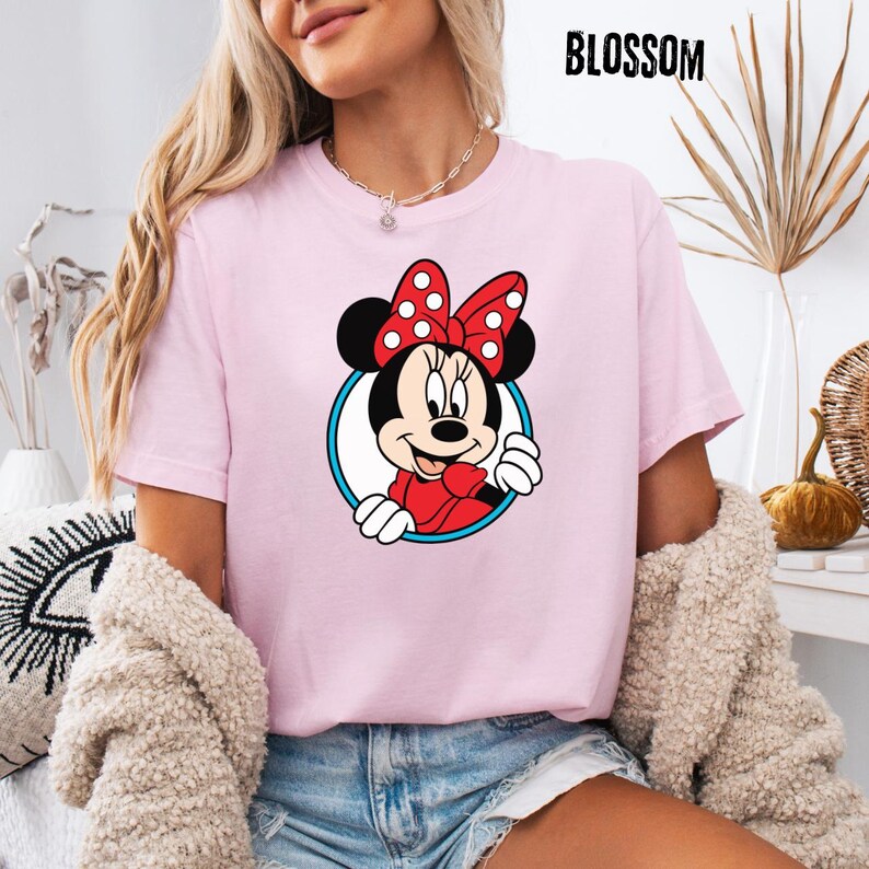 May include: A light pink t-shirt featuring a Minnie Mouse graphic. The cartoon character is framed in a blue and white circle, wearing a red bow with white polka dots. The word "BLOSSOM" is printed in black at the top.