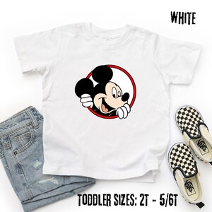 Pu&ograve; includere: T-shirt bianco per bambini piccoli con una grafica di Topolino sorridente all'interno di un cerchio rosso. La maglietta ha maniche corte e girocollo. Il testo "WHITE" &egrave; stampato in alto e "TODDLER SIZES: 2T - 5/6T" in basso.