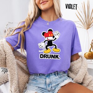 May include: A violet t-shirt featuring a cartoon mouse character with a red, black, and yellow design. The mouse is waving, and the word "DRUNK" is printed below. The word "VIOLET" is printed in the upper right corner.