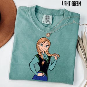May include: Light green t-shirt with a cartoon illustration of a character with blonde braids, wearing a blue and black outfit. A silver necklace with a pendant is draped across the shirt. The text "LIGHT GREEN" is at the top.