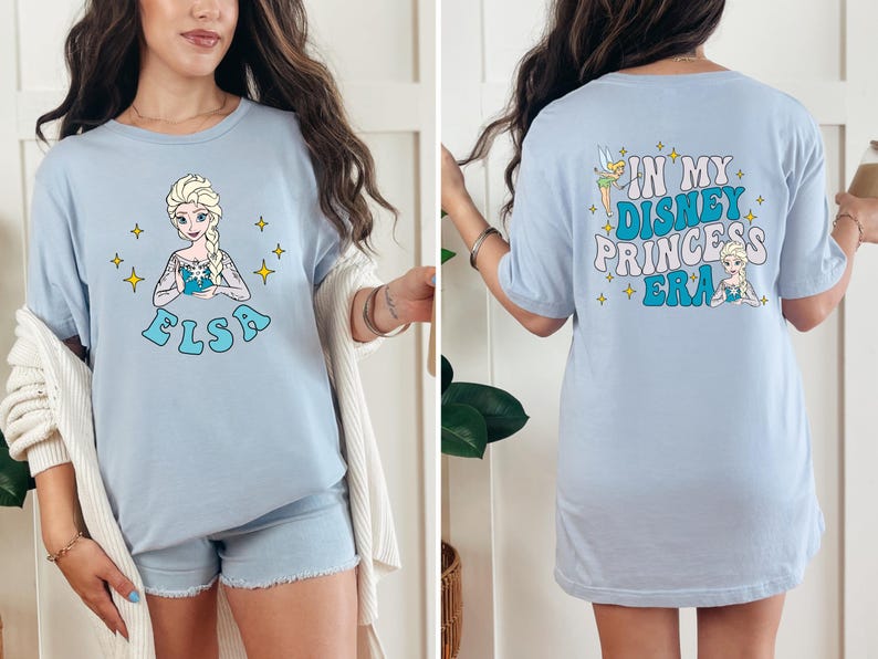 May include: A light blue t-shirt featuring a graphic of Elsa on the front with the name Elsa below. The back of the shirt displays the text In My Disney Princess Era with illustrations of Tinker Bell and Elsa, accented by small yellow star shapes.