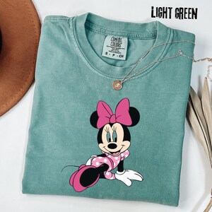 May include: Light green t-shirt featuring a Minnie Mouse graphic. The cartoon character is wearing a pink polka dot dress and bow. The shirt has a crew neck and short sleeves. A silver necklace with a pendant is also visible.