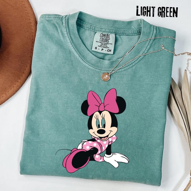 May include: Light green t-shirt featuring a Minnie Mouse graphic. The cartoon character is wearing a pink polka dot dress and bow. The shirt has a crew neck and short sleeves. A silver necklace with a pendant is also visible.