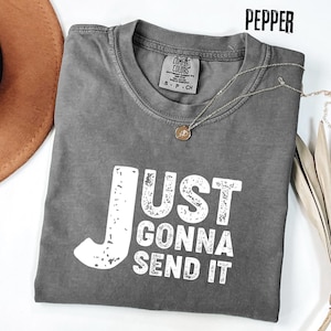 May include: A gray t-shirt with the text "JUST GONNA SEND IT" in white block letters. The shirt has a crew neck and short sleeves. A silver necklace with a small pendant is draped across the shirt. The shirt is folded on a white surface.