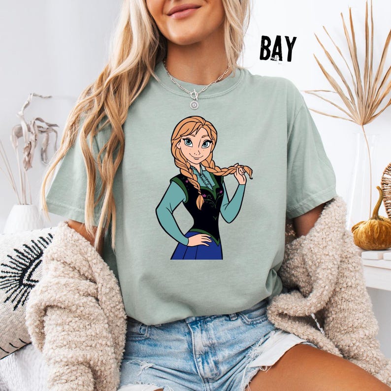 May include: A pale green t-shirt with a cartoon image of a character with blonde braids, wearing a black vest and blue dress. The word "BAY" is printed in black on the upper right side of the shirt.