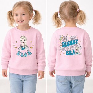 May include: A light pink long-sleeve sweatshirt featuring a graphic of Elsa on the front and the text IN MY DISNEY PRINCESS ERA on the back, accompanied by illustrations of Tinker Bell and Elsa, surrounded by small yellow star accents.