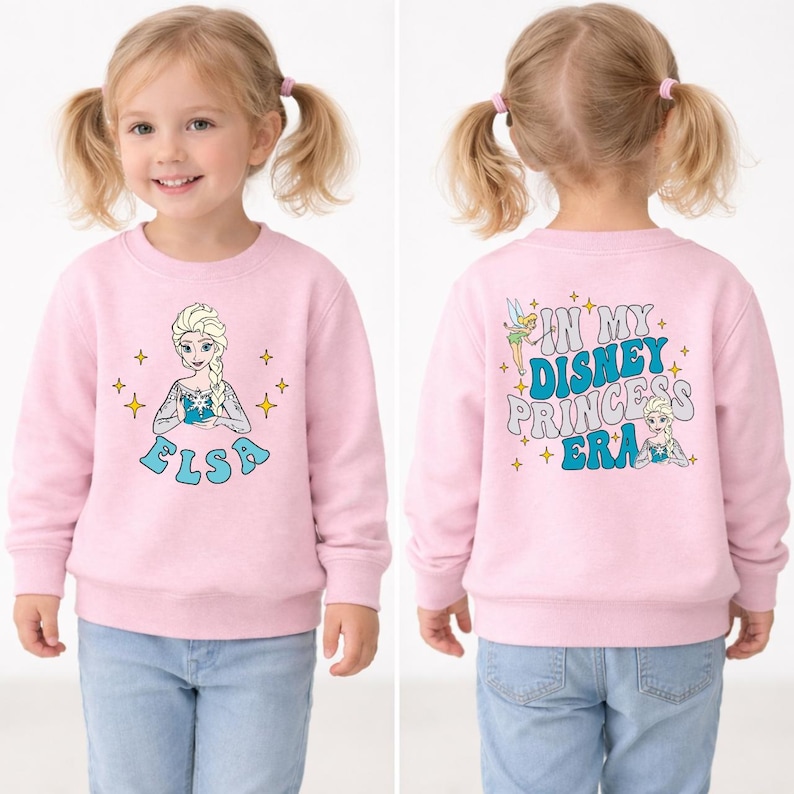 May include: A light pink long-sleeve sweatshirt featuring a graphic of Elsa on the front and the text IN MY DISNEY PRINCESS ERA on the back, accompanied by illustrations of Tinker Bell and Elsa, surrounded by small yellow star accents.